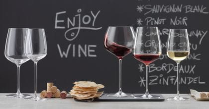 SPIEGELAU Willsberger Anniversary glasses on a marble sideboard. On the left an empty Bordeaux glass and white wine glass, in the middle a pile of corks and potato chips, on the right the filled Burgundy glass, filled red wine glass and filled white wine glass. In the background a blackboard with a menu and the words drink wine.<br/>