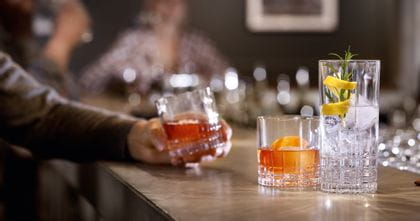 A bar counter with two SPIEGELAU Perfect Serve Collection glasses. The SOF glass is filled with an orange colored drink and an orange zest, the longdrink glass is filled with clear cocktail drink, ice cubes, rosemary and a lemon zest. In the background a hand, holding another filled SOF glass.<br/>