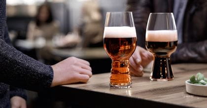 The SPIEGELAU Craft Beer Glasses IPA glass filled with IPA beer and the Stout glass filled with Stout beer standing on a wooden table. Behind them a hand reaching for the Stout glass.<br/>