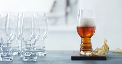A group of SPIEGELAU Craft Beer Glasses for IPA on a table. One standing on a wooden coaster, filled with IPA beer. Next to this glass are some potato chips.<br/>