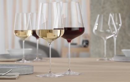 A SPIEGELAU Definition Bordeaux glass filled with red wine and a SPIEGELAU Definition white wine glass filled with white wine on a table. In the background are the same filled wine glasses again.<br/>