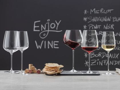 Copas SPIEGELAU Willsberger Anniversary sobre un aparador de mármol. A la izquierda una copa de Burdeos y una copa de vino blanco vacías, en el centro un montón de corchos y patatas fritas, a la derecha la copa de Borgoña llena, la copa de vino tinto llena y la copa de vino blanco llena. Al fondo una pizarra con un menú y las palabras beber vino.<br/>