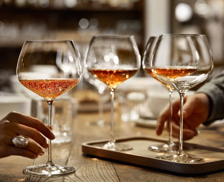 A hand holding the SPIEGELAU Burgundy glass of the series Willsberger Anniversary, filled with rosé colored sparkling wine. Further back on the wooden table, there are more glasses.<br/>
