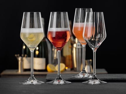 Cuatro copas de champán SPIEGELAU Lifestyle. Tres de ellas llenas de un cóctel de champán diferente, una de ellas vacía. Detrás, otra mesa con botellas de champán, limones y utensilios de bar.<br/>
