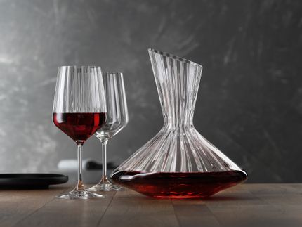 La jarra SPIEGELAU Lifestyle llena de vino tinto sobre una mesa. A la izquierda del decantador dos copas de vino tinto Lifestyle, una de ellas llena de vino tinto. En el borde izquierdo de la mesa un plato negro vacío.<br/>