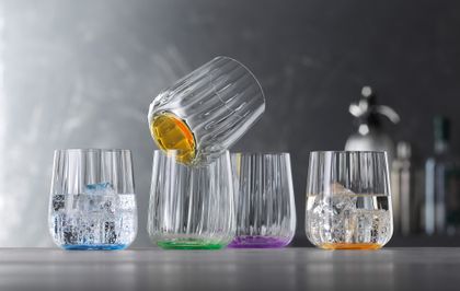 Las tazas SPIEGELAU Liefestyle en los colores océano, hoja, sol y lila están colocadas en fila. El vaso de color océano y sol está lleno de agua y cubitos de hielo.<br/>