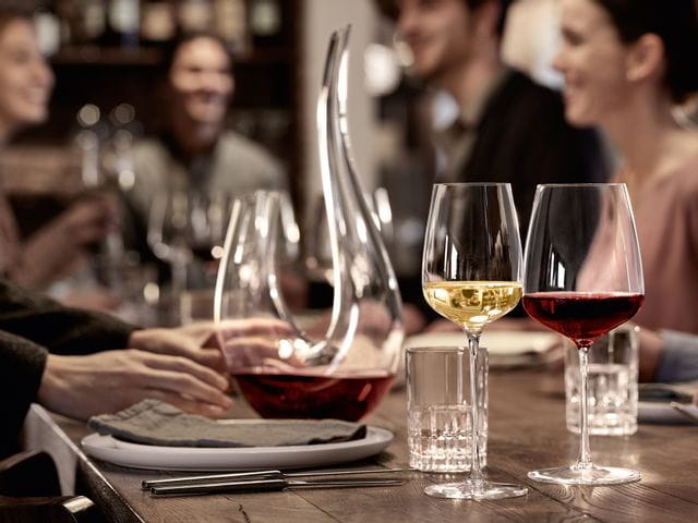 The filled white wine glass and the filled Bordeaux glass SPIEGELAU Willsberger Anniversary in front of the SPIEGELAU Perfect Serve Collection SOF glass and the filled SPIEGELAU Novo decanter. All of them on a dinner table surrounded by people.<br/>