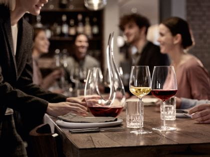 La copa de vino blanco rellena y la copa de Burdeos rellena SPIEGELAU Willsberger Anniversary delante de la copa SPIEGELAU Perfect Serve Collection SOF y la jarra rellena SPIEGELAU Novo. Todos ellos en una mesa rodeada de gente.<br/>