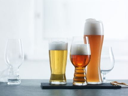 Cuatro vasos SPIEGELAU Beer Classics en fila, uno vacío y el resto llenos con diferentes cervezas. Los tres vasos llenos están colocados en una bandeja negra sobre un fondo claro.<br/>