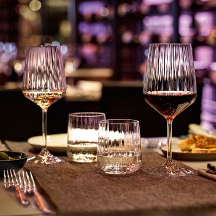 The SPIEGELAU Lifestyle series on a restaurant table in a cosy evening atmosphere. Purple light shines through the smooth wave structure of the filled white wine glass, red wine glass, water tumblers and carafe.<br/>