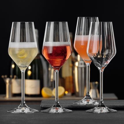 Four SPIEGELAU Lifestyle Champagne glasses. Three of them filled with a different Champagne cocktail, one of them empty. Behind them another table with Champagne bottles, lemons and bar equipment.<br/>