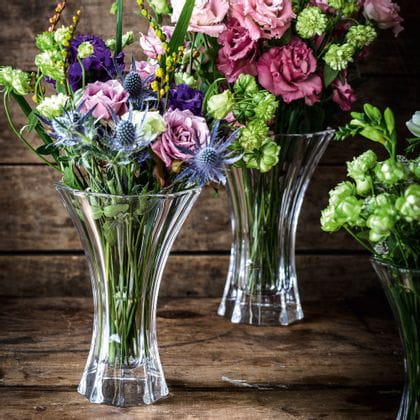 The crystal vases NACHTMANN Saphir, filled with colorful flower bouquets in red, green, violet and blue on a wooden sideboard.<br/>