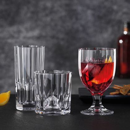 The NACHTMANN series Aspen on a black table. The goblet to the right is filled with a red cocktail, ice cubes and an orange slice. The tumbler and long drink glass to the left are empty.